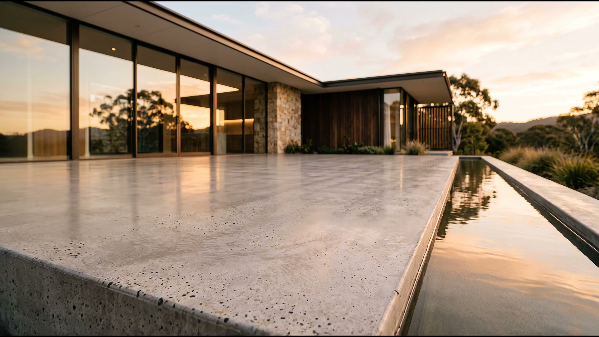 Burnished concrete terrace of a contemporary house at golden hour, looking across the slab to glass and stone